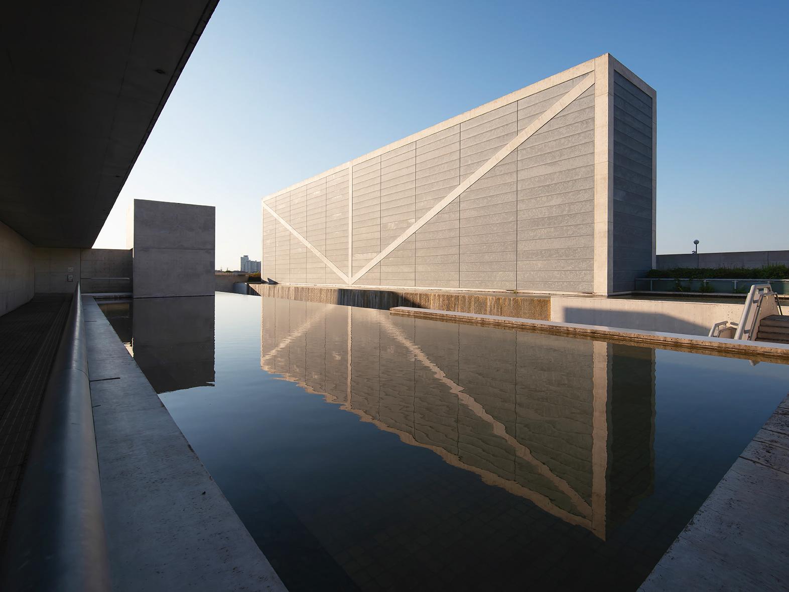 Stone-clad building with a diagonal structural brace reflected in a still pool